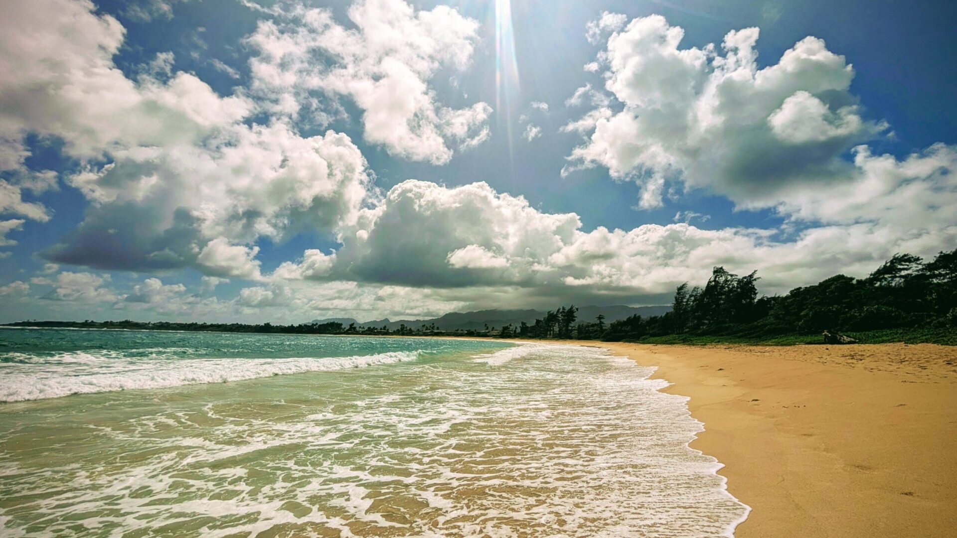 Sunny day at Hauʻula Beach on Oʻahu's North Shore with foamy waves, golden sand, and scattered clouds under a bright blue sky