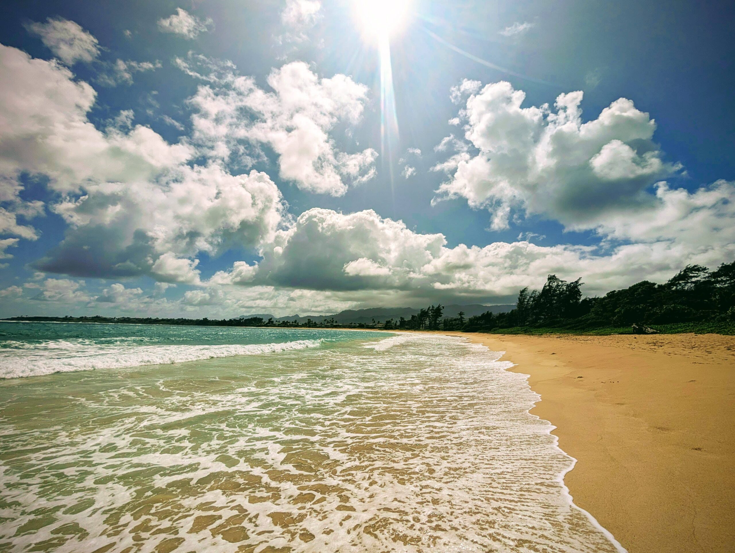 Sunny day at Hauʻula Beach on Oʻahu's North Shore with foamy waves, golden sand, and scattered clouds under a bright blue sky