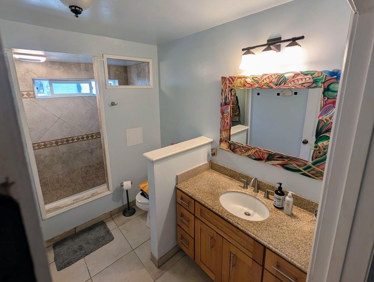Clean and tropical bathroom at Hauʻula Hideaway featuring a walk-in tiled shower and artistic mirror