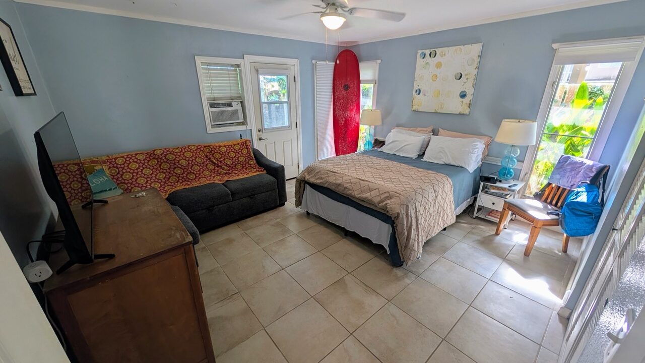 Bright guest suite with queen bed, ceiling fan, sleeper sofa, TV, and surfboard decor in Hauʻula Hideaway